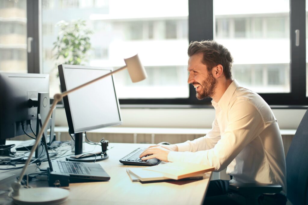 pexels photo 840996 840996 A man smiling while working at an office desk with a computer and natural daylight streaming in through large windows.